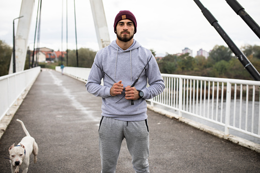 A handsome athlete is posing for a photo standing on a suspension bridge with a speed rope around his shoulders and his dog next to him