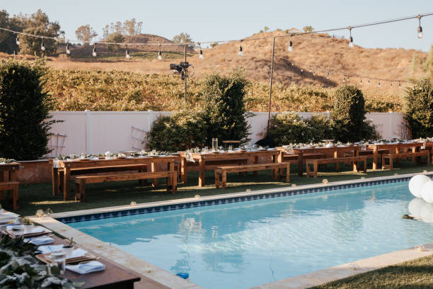 istock Backyard wedding setting - wooden tables with bohemian decor around swimming pool. 1283746045