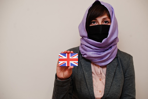 Portrait of young muslim woman wearing formal wear, protect face mask and hijab head scarf, hold United Kingdom flag card against isolated background. Coronavirus country concept.