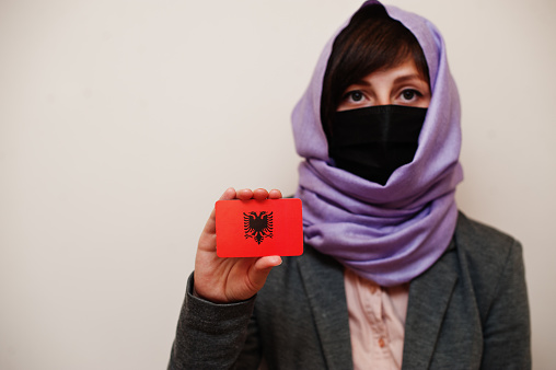 Portrait of young muslim woman wearing formal wear, protect face mask and hijab head scarf, hold Albania flag card against isolated background. Coronavirus country concept.