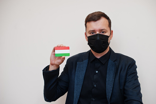 European man wear black formal and protect face mask, hold Hungary flag card isolated on white background. Europe coronavirus Covid country concept.