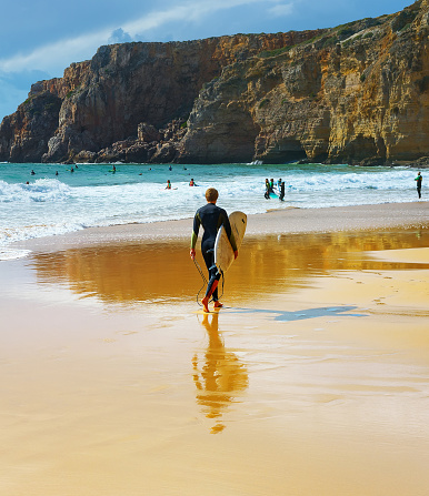SAGRES, PORTUGAL - OCTOBER 30, 2018: Man walking by sandy beach with surfboard. ALgarve is a famous surfing destination in Portugal
