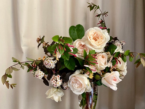 Closeup photo of a beautiful bouquet of pastel cream and pink roses, yellow and pink honeysuckle and Hawthorne blossom in a glass vase on a rustic wooden table in front of a pale curtain