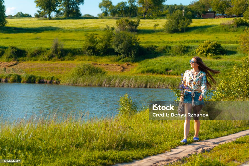 Beautiful young brunette slender woman with long hair in jeens shorts and loose flower blouse walking past the lake on the ground road, countriside 35-39 Years Stock Photo