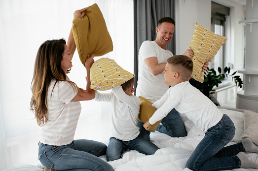 Young parents with sons fighting pillows on the bed. Happy mother and father with boys having fun at home.