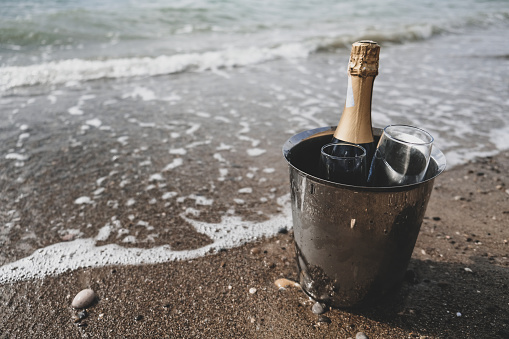 Ocean waves chilling drinks for tourists, luxury vacation for two. Ice bucket with champagne bottle and glasses on a beach surrounded by sea. Anniversary day, romantic holiday or honeymoon together.