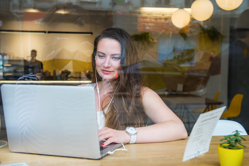 View through the window with city reflection of smiling businesswoman in headset working on laptop sitting in cafe, coworking. Remote work, social networks, blogging. Telecommuting concept. Freelancer.