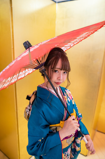 Young woman in ‘furisode’ kimono holding paper umbrella and standing in front of golden ‘Byobu’ folding screen for ‘Seijin Shiki’ coming-of-age ceremony