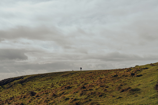 Unidentifiable man and a dog walking on the horizon line on top of a hill in Mendip Hills, UK, against the sky.