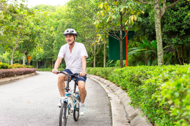 Senior Asian men cycling road bike on a forest road in morning male cyclist with safety helmet riding a foldable bicyle in beautiful public park near a hill, as he padeling downhill man 60s bicycle stock pictures, royalty-free photos & images