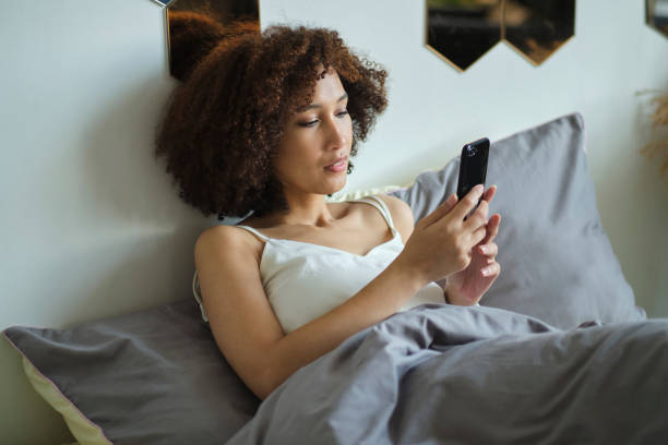 istock Calm beautiful african woman in pajamas using smartphone while lying in bed isolated 1283810175