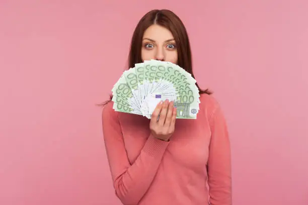 Amazed happy woman with brown hair in pink sweater hiding face behind fan of euro banknotes, interest-free cash withdrawal. Indoor studio shot isolated on pink background