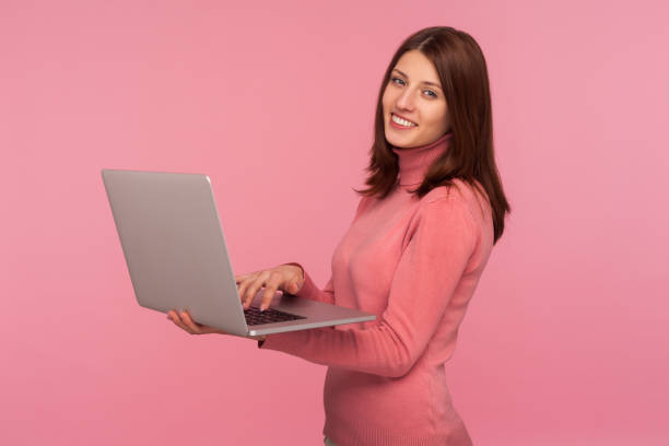 femme positive heureuse avec des cheveux bruns dans le chandail rose travaillant sur l’ordinateur portatif et regardant l’appareil-photo avec le sourire denté, appréciant le travail à distance. - women computer home interior brown hair photos et images de collection