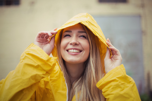 Closeup view of a cute young, woman with a hood on her head. Her smile and the yellow raincoat that she is wearing is making us love autumn even more.