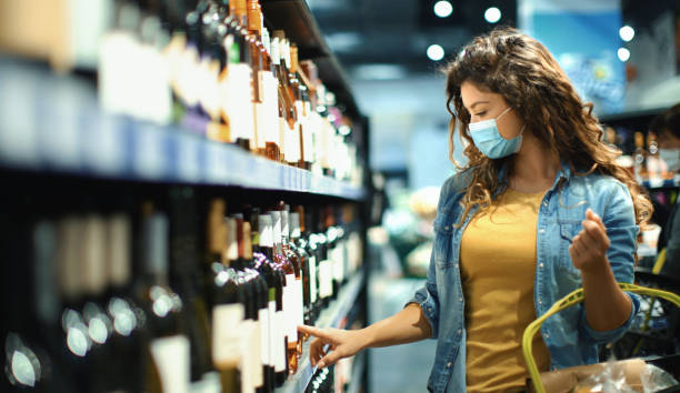 Young woman buying some wine at a supermarket during coronavirus pandemic Closeup side view of a late 20's woman going grocery shopping during coronavirus pandemic. She's choosing some wine for tonight's dinner. alcohol shop stock pictures, royalty-free photos & images