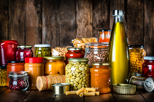 Front view of a group of non-perishable food shot on rustic wooden table. The composition includes tins, sauces, dried legumes, crackers, pasta, preserves, peanut butter, cooking oil among others. High resolution 42Mp studio digital capture taken with Sony A7rII and Sony FE 90mm f2.8 macro G OSS lens