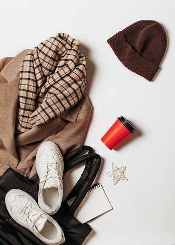 Fashion flat lay with beige or brown coat casual, black bag, cap, plaid scarf, sneakers and red caup of coffee on white background, top view, copy space,
