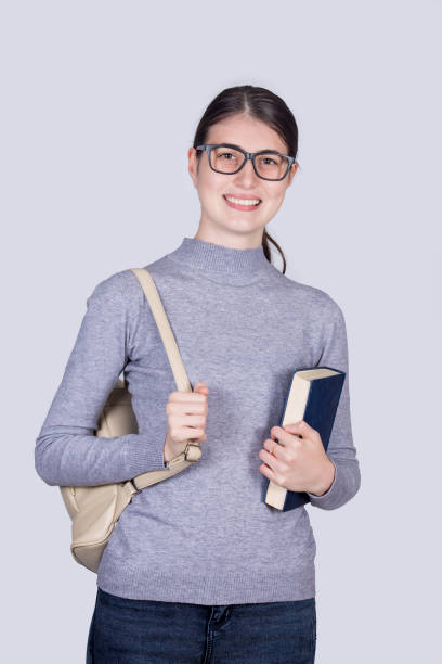 istock Confident student girl  smiling holding one books and carrying her backpack. Cheerful student girl  with positive face expression isolated on white background. 1283848144