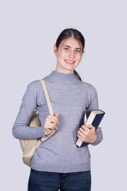 istock Confident student girl  smiling holding one books and carrying her backpack. Cheerful student girl  with positive face expression isolated on white background. 1283848306