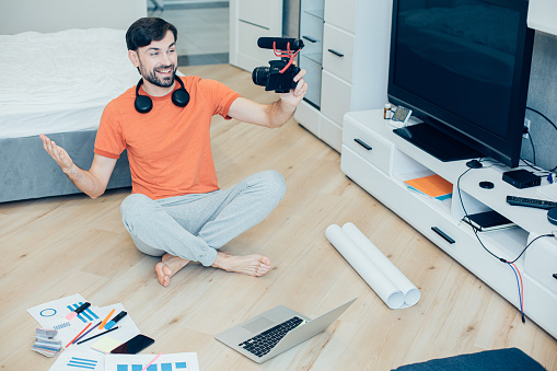 Mirthful Caucasian male blogger sitting on the floor of a bedroom and smiling while gesturing and recording himself