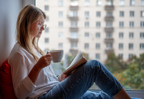 Young woman with long red hair in glasses in a white shirt sitting on the windowsill at home reading book drinking coffee