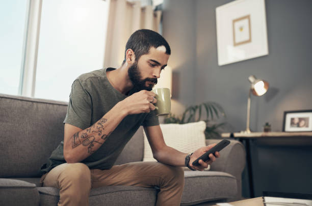 Connect to all things current with the ease of technology Shot of a young man having coffee and using a smartphone on the sofa at home bachelor stock pictures, royalty-free photos & images