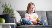 Adorable child girl 8 years old sits on a gray sofa with a green apple and a TV remote control in her hands rest and entertainment at home