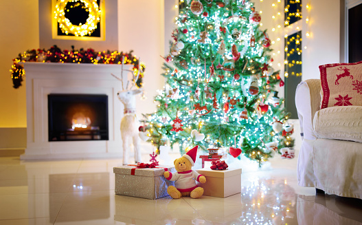 Christmas tree in decorated living room. Family home winter season decoration. Present and gift boxes on Xmas eve. Fireplace with garlands, ornaments and lights. Couch with knitted reindeer cushions.