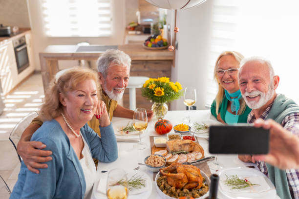 Senior friends taking a selfie while having Thanksgiving dinner Senior people having fun while celebrating Thanksgiving together at home over traditional dinner, taking a selfie using smart phone thanksgiving holiday covid stock pictures, royalty-free photos & images