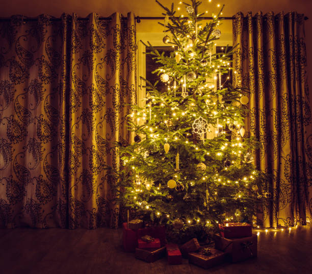 istock Christmas tree with warm yellow string lights and white icicles and snowflake shape ornaments. Red packed presents under the tree. 1283868432
