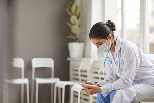 Young nurse in mask and in white coat using her mobile phone during her break sitting on chair at the corridor