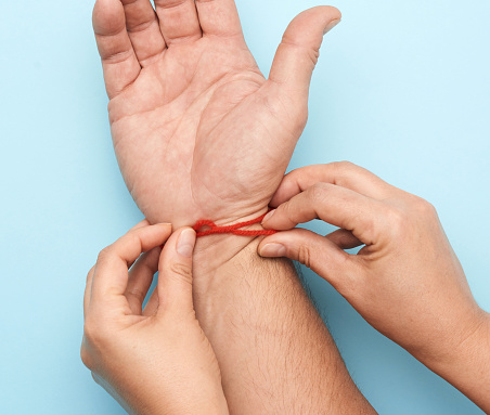 two female hands tie a red woolen thread on the wrist of a male hand, blue background, top view