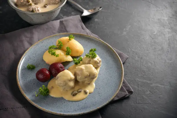 Boiled meatballs in Germany called Koenigsberger Klopse in a white bechamel sauce with capers, potatoes and beetroot on a blue plate, dark gray napkin and slate background with copy space, selected focus, narrow depth of field