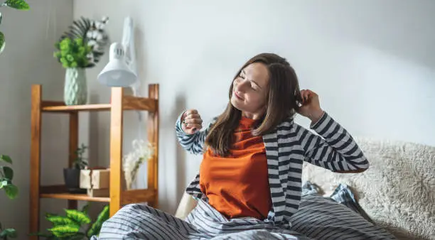 Happy girl in pajamas is sitting on the bed in the sunny bedroom and stretches after waking up. Concept of a nice start to the day and good mood.