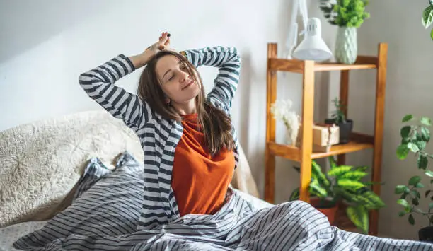 Happy girl in pajamas is sitting on the bed in the sunny bedroom and stretches after waking up. Concept of a nice start to the day and good mood.