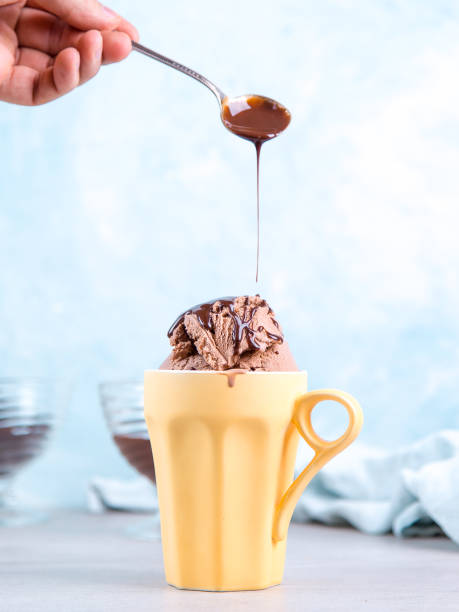 istock Chocolate brown ice cream in a regular yellow mug, on a blue background, the hand pours from a spoon topping. Unusual serve of dessert, copy space. 1283885048