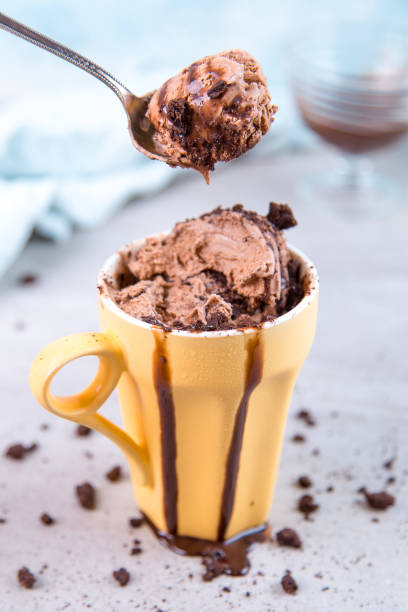 istock Chocolate brown ice cream in a regular yellow mug and spoon, on a blue background, with topping and crumbs. Unusual serve of dessert, copy space. 1283885069