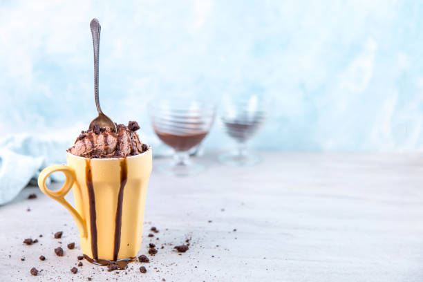 istock Chocolate brown ice cream in a regular yellow mug and spoon, on a blue background, with topping and crumbs. Unusual serve of dessert, copy space. 1283885071