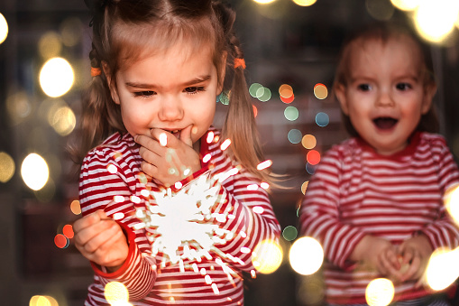 Family holidays at home. Two happy kids, toddler boy and cute small girl holding burning sparkler and smile happily over defocused festive background, indoor emotional lifestyle