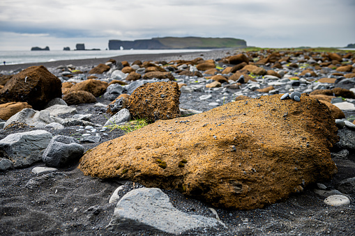 Vik, Iceland black sand beach ocean horizon with rock formation arch at Dyrholaey, Kirkjufjara and foreground closeup of brown stones