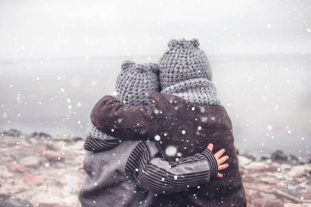 Girl in knitted grey hat hugging her frozen smaller brother Little pretty girl in knitted grey hat hugging her frozen smaller brother during snowfall in winter, white cold weather, winter outside lifestyle close-up portrait hood clothing stock pictures, royalty-free photos & images