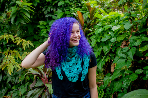 Portrait of young lady with purple hair wearing a blue crochet shawl. Purple haired girl touching her hair and smiling.