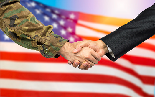 American soldier in uniform and civil man in suit shaking hands with adequate national flag on background