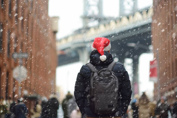 middle aged man tourist in a santa claus hat is walking during a snowfall near manhattan bridge in new york on a snowy christmas eve. winter xmas holidays in nyc. - backpack one mature man only only mature men one man only imagens e fotografias de stock