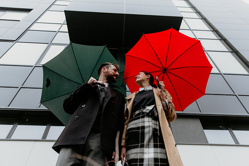 Standing still. Both wearing their elegant business wear. One holding the red and other dark green umbrella open. Looking at each other with a big smile on their face. The business building behind them.