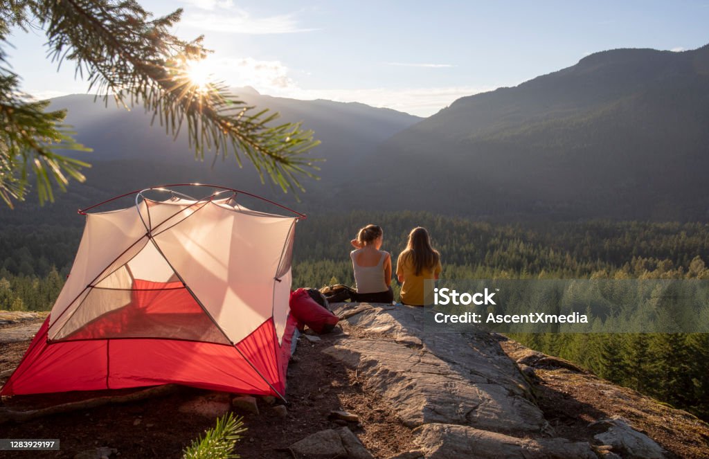 Young women watch the sunset on a mountain ledge campsite Their tent is illuminated in the sunlight Camping Stock Photo