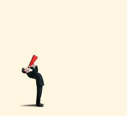 A businessman bends over backward as he shouts into a red megaphone isolated against a yellow background.