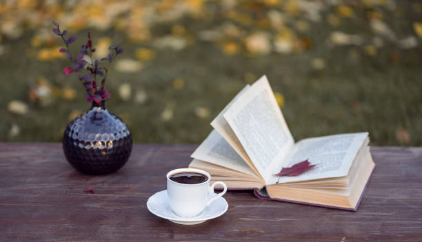 istock Little white cup of espresso coffee, opened book with maple leaf inside, vase with Berberis branches on rustic wooden table in the garden full of fallen leaves at autumn day 1283942939