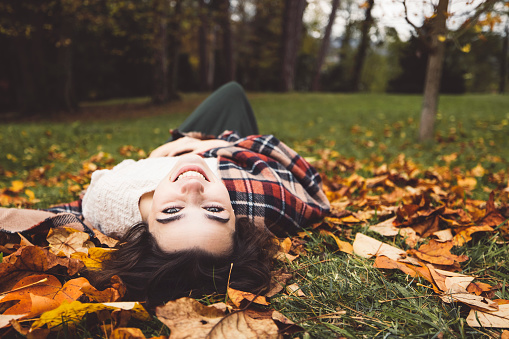 Smiling caucasian young woman laying on the grass with autumn leaves all around.