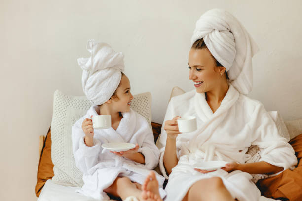 feliz familia madre y hija pequeña en albornoz y toallas descansar y relajarse con una taza de café en la cama - bathrobe women cup towel fotografías e imágenes de stock